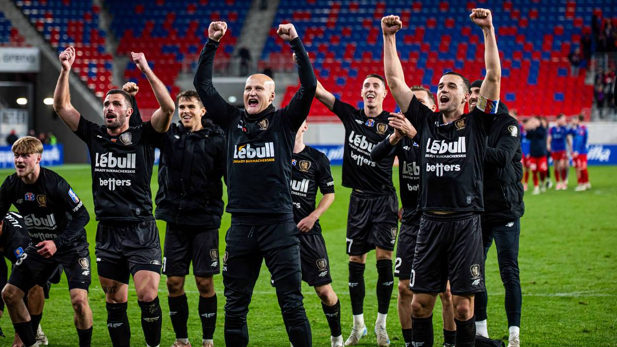 Piłkarze w czarnych strojach świętują na boisku z podniesionymi rękami.