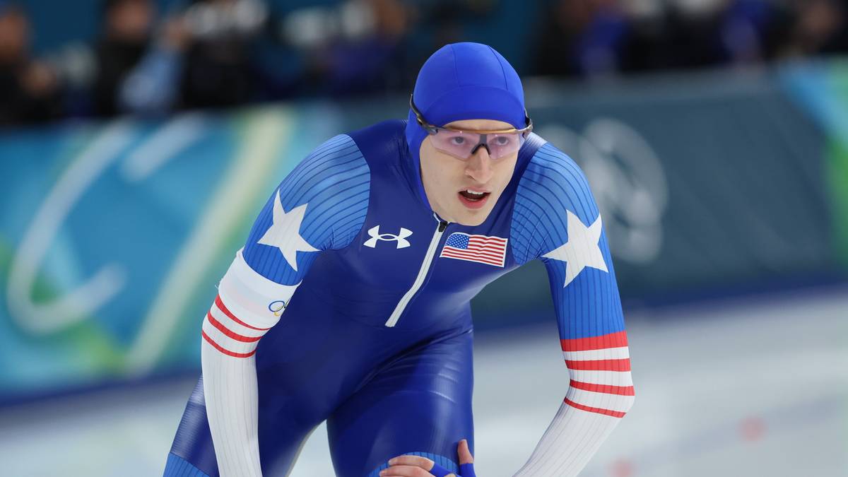 Łyżwiarz szybki w niebieskim kombinezonie z białymi gwiazdami i czerwono-białymi pasami na rękawach i nogawkach, noszący okulary ochronne i niebieską czapkę, w trakcie biegu na lodowisku.