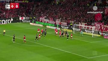 Fragment meczu piłki nożnej FSV Mainz - SC Freiburg. Gracze w czerwonych i czarnych strojach na boisku, trybuny wypełnione kibicami, wynik 0:0.