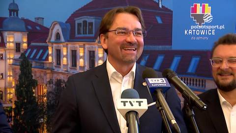 Sebastian Świderski, prezes Polskiego Związku Piłki Siatkowej, uśmiecha się podczas konferencji prasowej z mikrofonami przed nim i budynkami w tle.