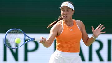 Tenisistka w pomarańczowej koszulce i białej sp&oacute;dniczce wykonuje uderzenie forhendowe podczas meczu.