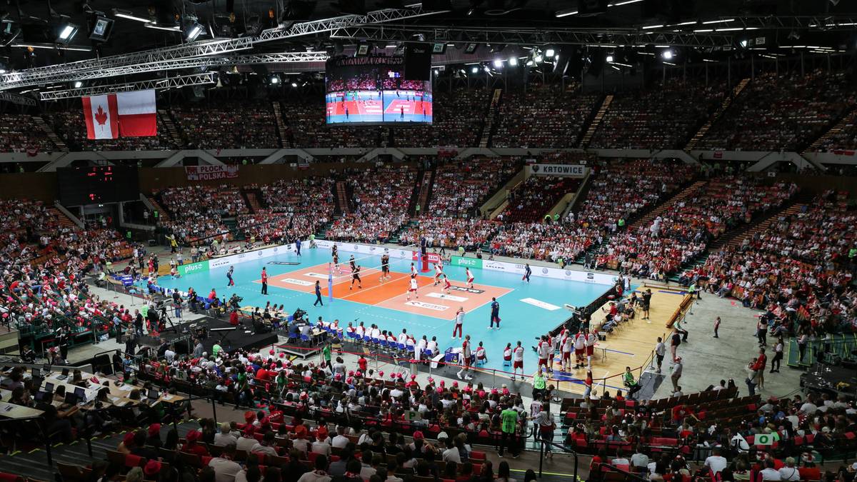 Widok na halę sportową podczas meczu siatkówki, z zawodnikami na boisku i kibicami na trybunach.