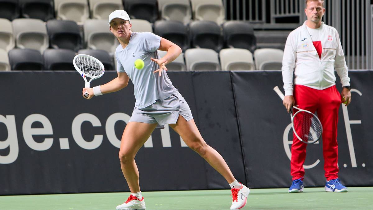 Tenisistka w szarej koszulce i spódnicy, podczas meczu, uderza piłkę rakietą. Za nią stoi mężczyzna w białej bluzie i czerwonych spodniach.