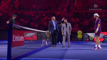 Dwóch mężczyzn w garniturach stoi przy siatce tenisowej, jeden z nich podaje rękę drugiemu. Po lewej stronie, za siatką, widać gracza w czapce i koszulce. Po prawej stronie, na korcie, stoi drugi gracz w sportowym stroju.