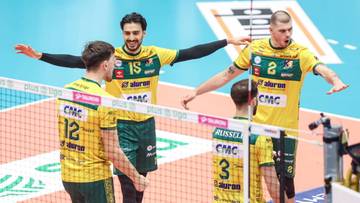 Siatkarze w ż&oacute;łtych koszulkach z zielonymi detalami, z numerami na plecach, stoją przy siatce, rozmawiają ze sobą.