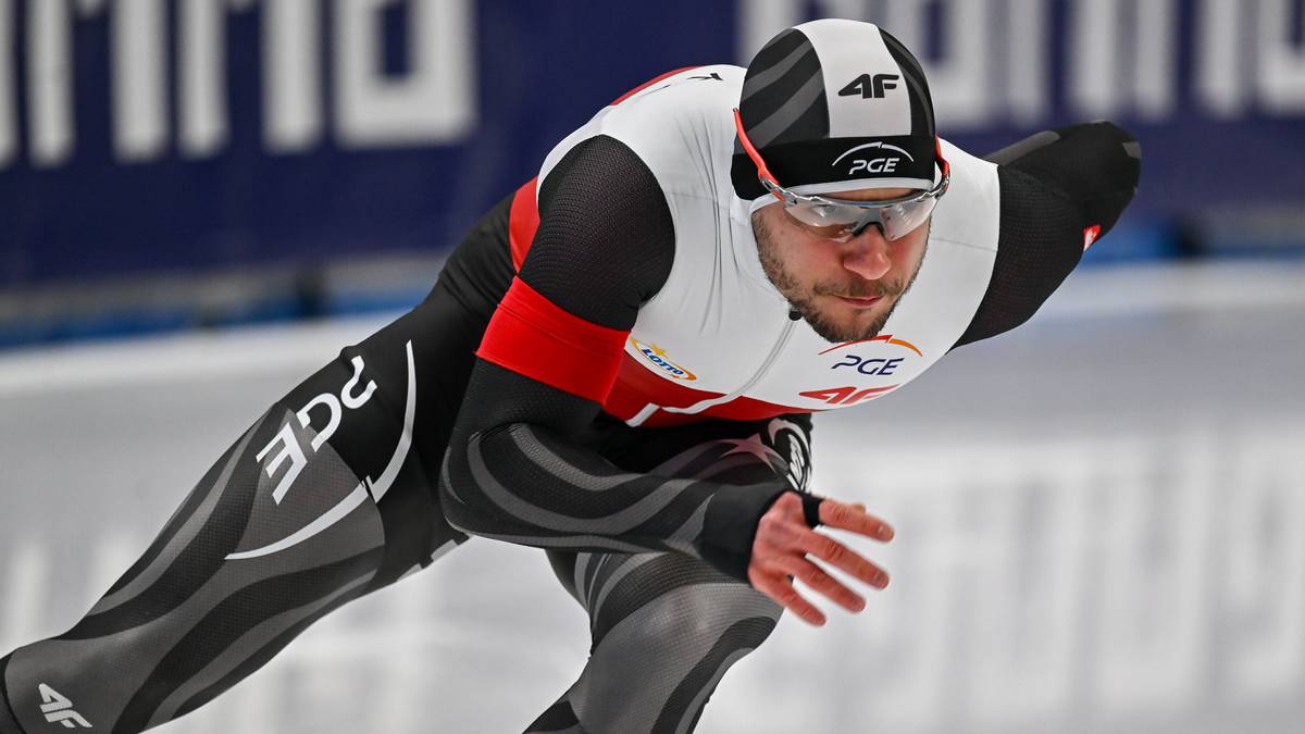 Zawodnik łyżwiarstwa szybkiego w kombinezonie i kasku z goglami, pochylony podczas jazdy na lodzie.