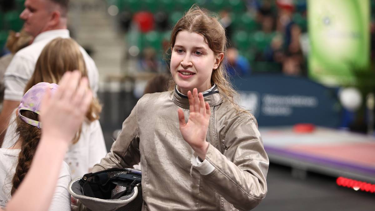 Uśmiechnięta młoda kobieta w stroju szermierczym, z ręką uniesioną w geście powitania lub radości, trzyma w drugiej ręce kask.