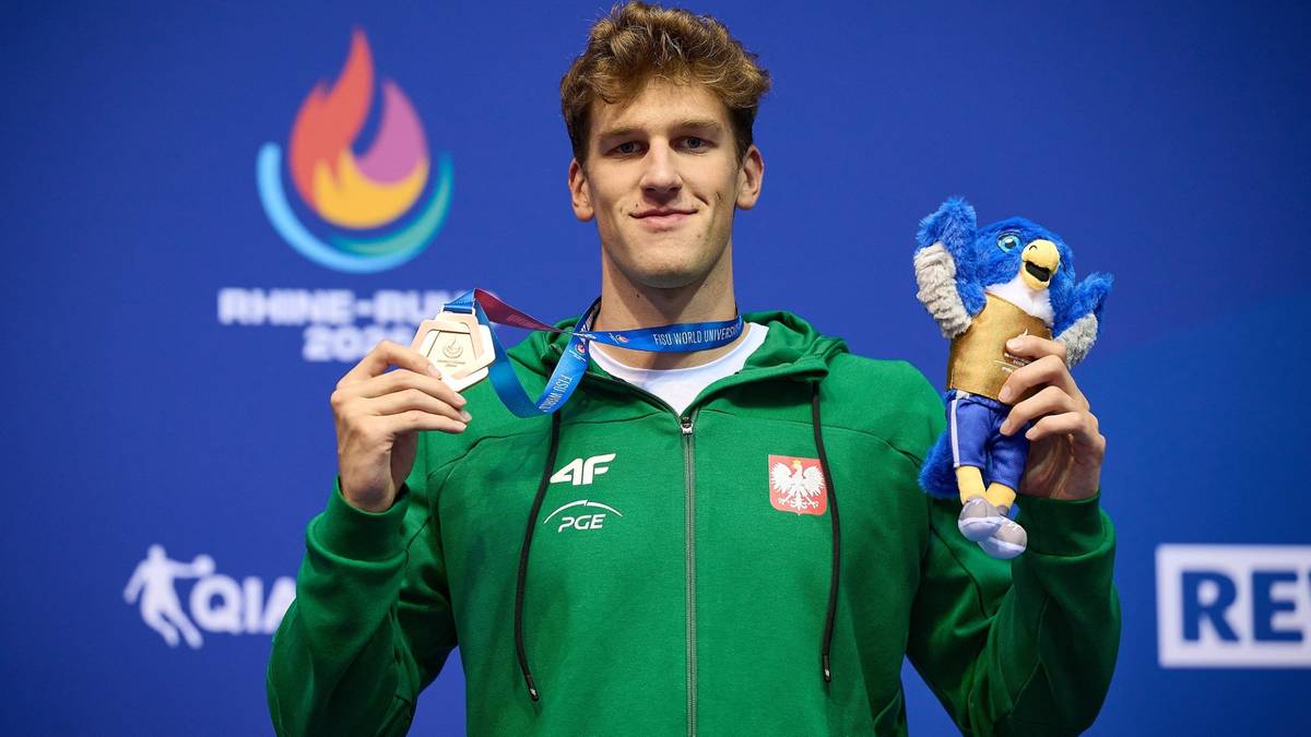 Młody mężczyzna w zielonej bluzie z kapturem z polskim orłem trzymający medal i pluszową zabawkę.