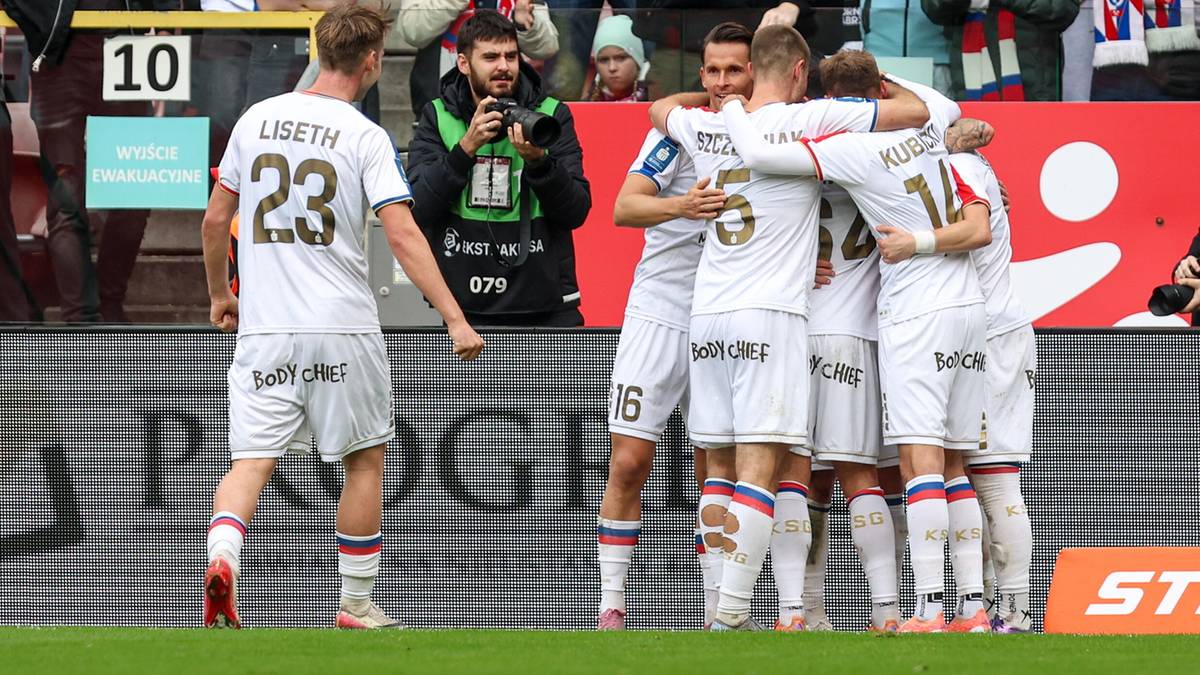 Piłkarze Górnika Zabrze w białych strojach świętują zdobycie bramki, stojąc w grupie na boisku. Jeden z nich odwrócony tyłem, inni patrzą w jego kierunku. W tle widać kibiców na trybunach i fragment stadionu.