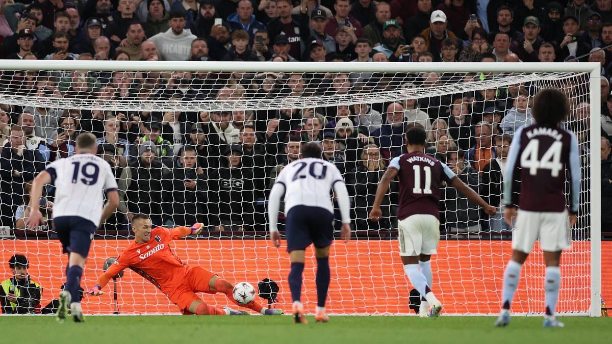 Piłkarz w pomarańczowym stroju bramkarza broni rzut karny wykonywany przez zawodnika w białej koszulce. W tle widoczni są inni piłkarze i trybuny zapełnione kibicami.