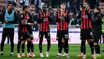 Pięciu piłkarzy AC Milan w czarno-czerwonych strojach stoi na boisku, patrząc w stronę kibic&oacute;w na trybunach.