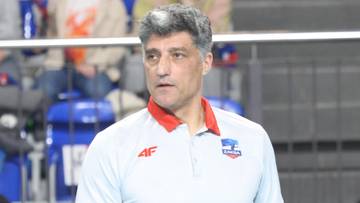 Trener siatk&oacute;wki w jasnej koszulce polo z kołnierzykiem w kolorze czerwonym, patrzący przed siebie.