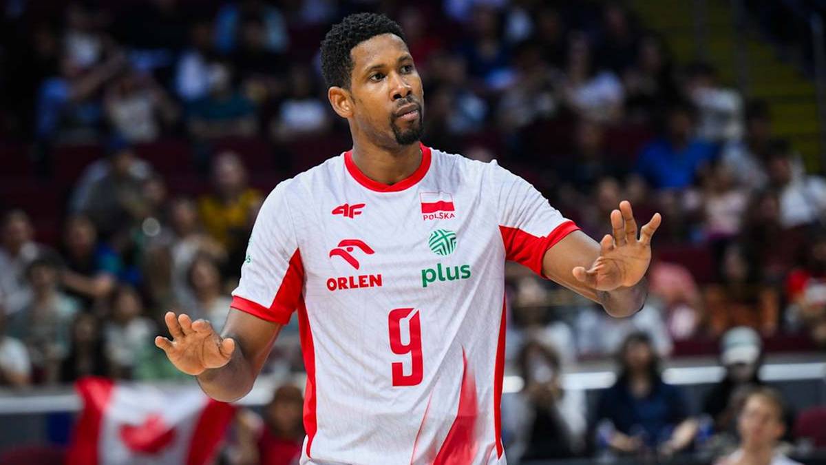 Siatkarz w białej koszulce z czerwonymi akcentami, z numerem 9 na piersi, pozycjonuje się na boisku, patrząc w stronę przeciwnika. W tle widać trybuny wypełnione widzami.