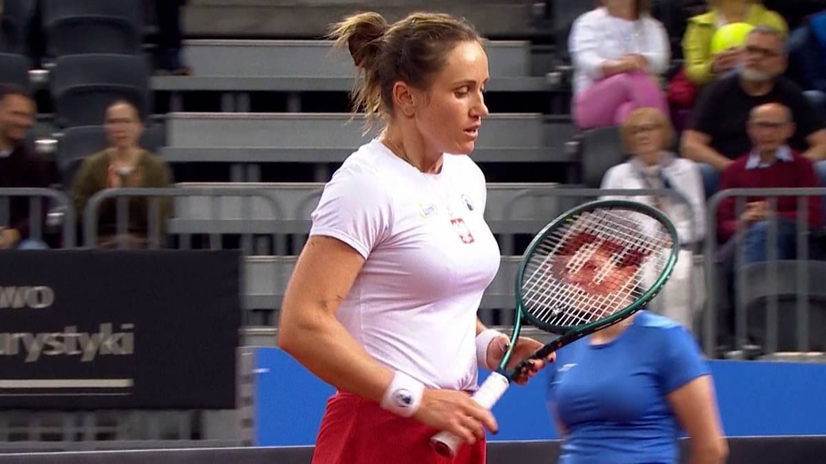 Tenisistka Katarzyna Kawa podczas meczu, trzymająca rakietę tenisową, w białej koszulce i czerwonych spodenkach.