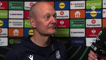 Portret mężczyzny w średnim wieku z kr&oacute;tkimi, siwymi włosami, patrzącego w bok. Tło stanowi ściana z logo sponsor&oacute;w.