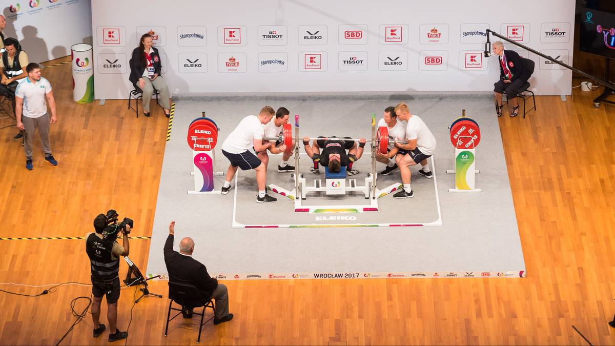 Podczas zawodów w trójboju siłowym na The World Games 2025, czterech mężczyzn w białych koszulkach i czarnych spodenkach podnosi sztangę na specjalistycznej ławce. Trzech z nich napina mięśnie, podczas gdy czwarty stoi z boku. Na widowni siedzą ludzie, a obok sceny znajduje się sprzęt filmowy.