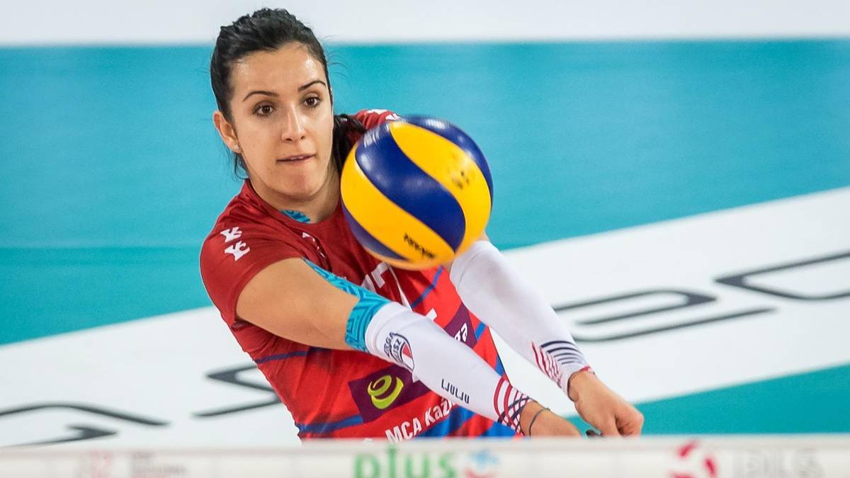 Siatkarka w czerwonym stroju z niebieskimi i żółtymi detalami odbija piłkę do siatkówki.