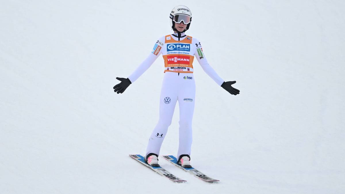 Skoczkini narciarska w białym stroju, z nartami na nogach, z rękami rozłożonymi na boki.