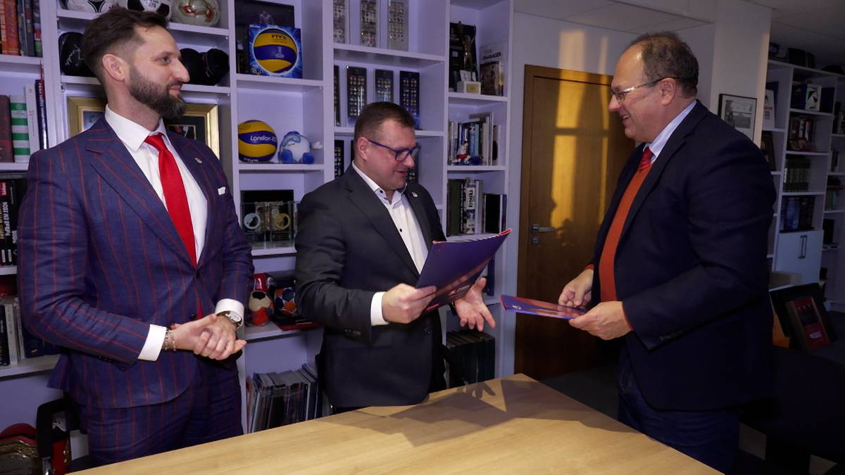 Trzech mężczyzn w garniturach podczas formalnego spotkania, jeden z nich podaje dokument.