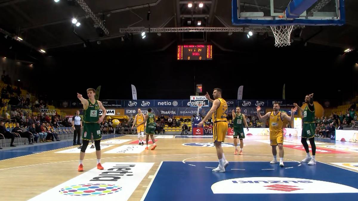 Mecz koszyk&oacute;wki z udziałem zawodnik&oacute;w w zielonych i ż&oacute;łtych strojach na parkiecie hali sportowej.