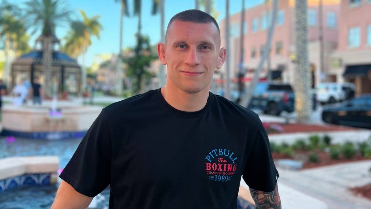 Mężczyzna w czarnej koszulce z logo Pitbull Boxing, pozuje do zdjęcia na tle basenu i palm.