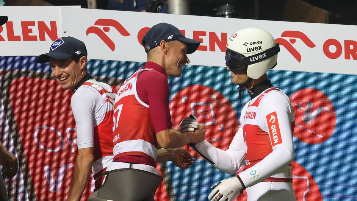 Trzech polskich skoczk&oacute;w narciarskich w białych i czerwonych strojach sportowych, jeden w kasku, gratuluje sobie nawzajem po zawodach.