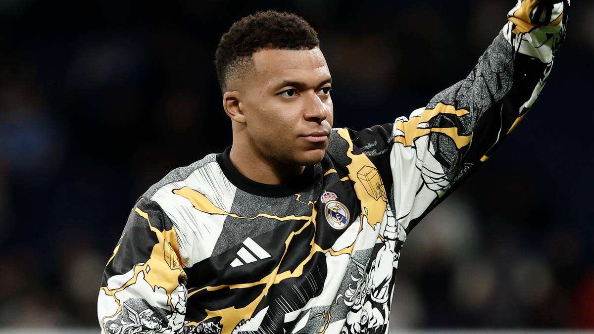 Piłkarz Kylian Mbappe w bluzie treningowej z charakterystycznym wzorem, macha do publiczności na stadionie.