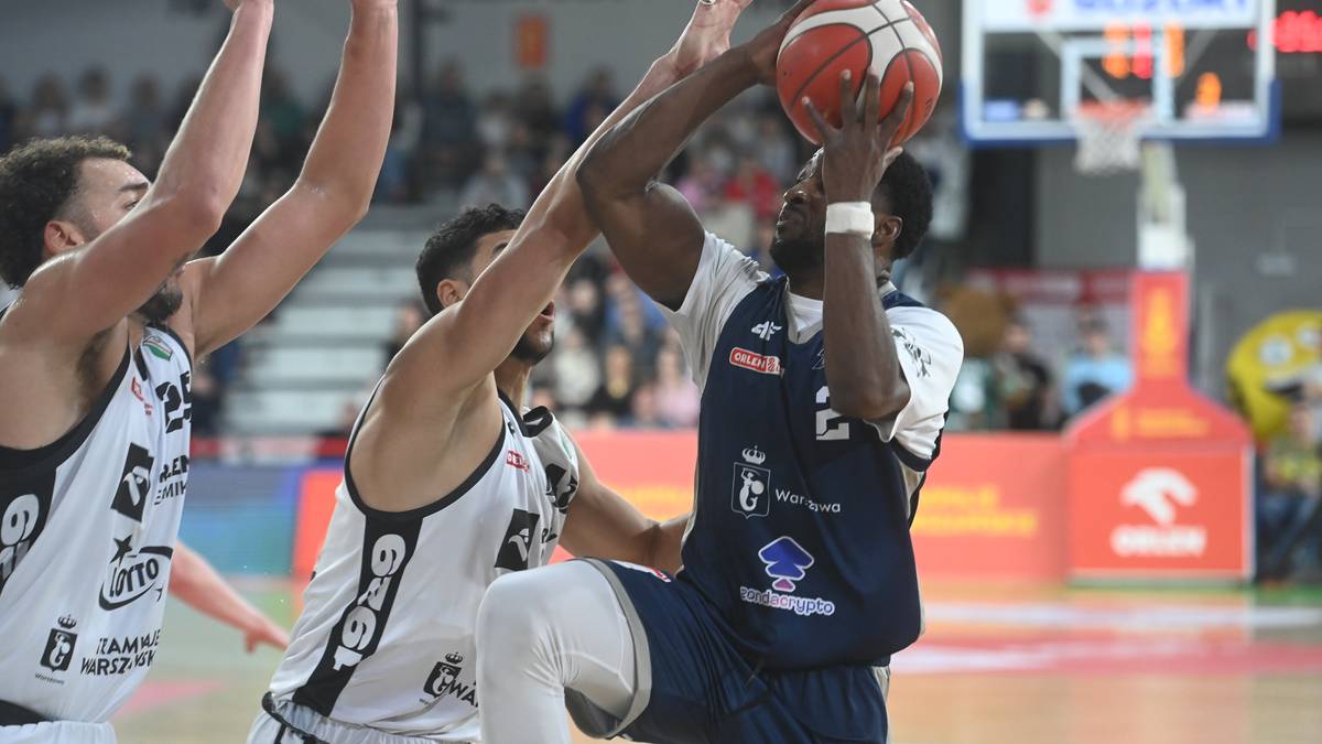 Koszykarz w niebieskim stroju z piłką w rękach, próbujący wykonać rzut, podczas gdy dwaj gracze w białych strojach próbują go powstrzymać.