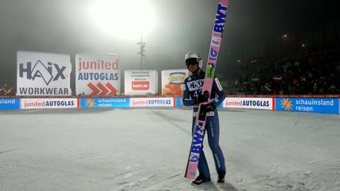 Skoczek narciarski w kombinezonie stoi na śniegu, trzymając narty pionowo. W tle widać reklamy i trybuny wypełnione widzami.