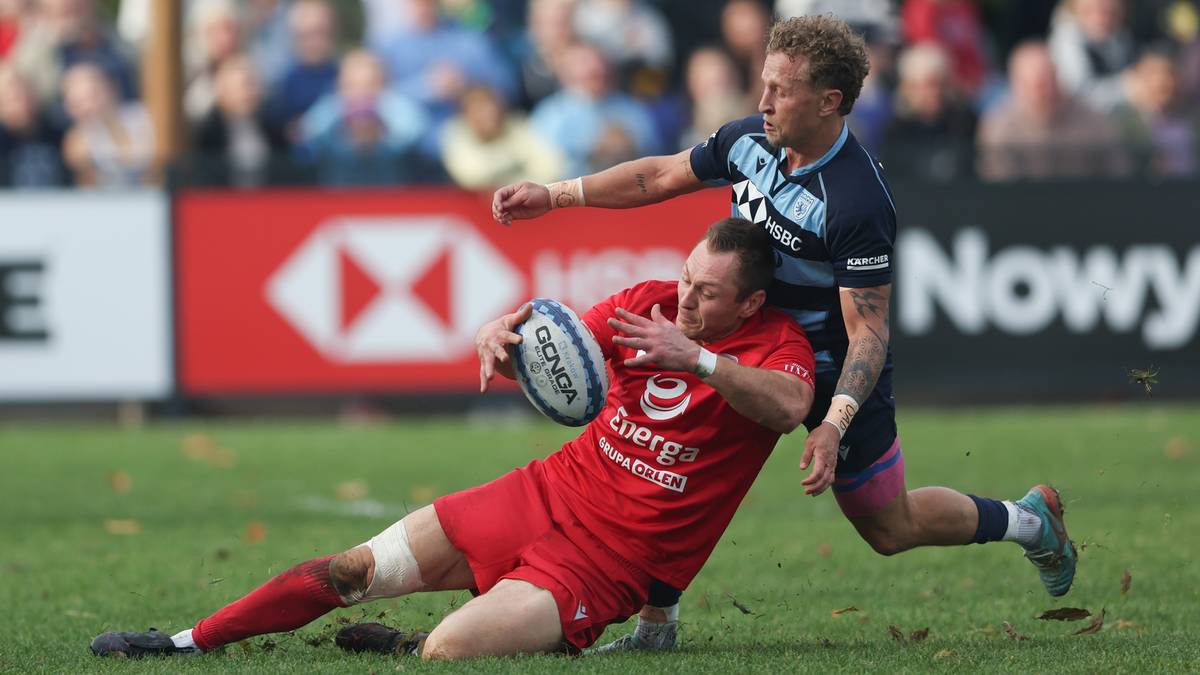 Gracz w czerwonym stroju z piłką do rugby próbuje przedostać się przez obronę przeciwnika, który próbuje go powstrzymać.