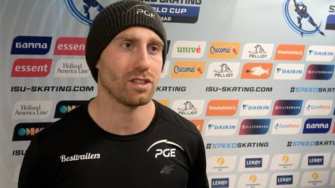 Damian Żurek, zawodnik speed skatingu, w czapce i bluzie z logo PGE, stoi przed ścianą z logotypami sponsor&oacute;w.