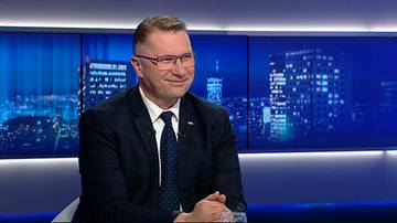 Przemysław Czarnek w ciemnym garniturze i krawacie, siedzi w studiu telewizyjnym z niebieskim tłem przedstawiającym nocne miasto. Uśmiecha się.