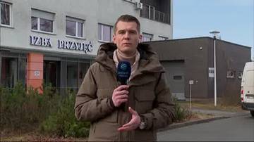 Dziennikarz w kurtce trzyma mikrofon na tle budynku szpitala z napisem "IZBA PRZYJĘĆ".
