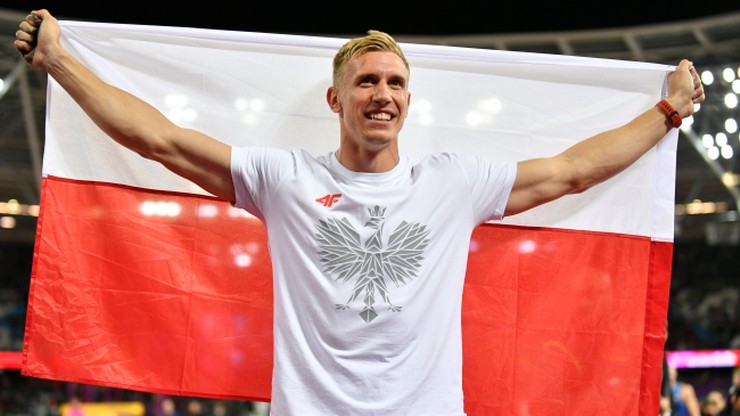 Uśmiechnięty biały mężczyzna z blond włosami, ubrany w białą koszulkę z orłem, trzyma rozłożoną polską flagę za jej końce nad ramionami.