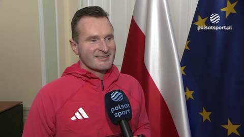 Maciej Maciusiak w czerwonej bluzie sportowej, trzymający mikrofon Polsat Sport, z polską flagą i flagą Unii Europejskiej w tle.