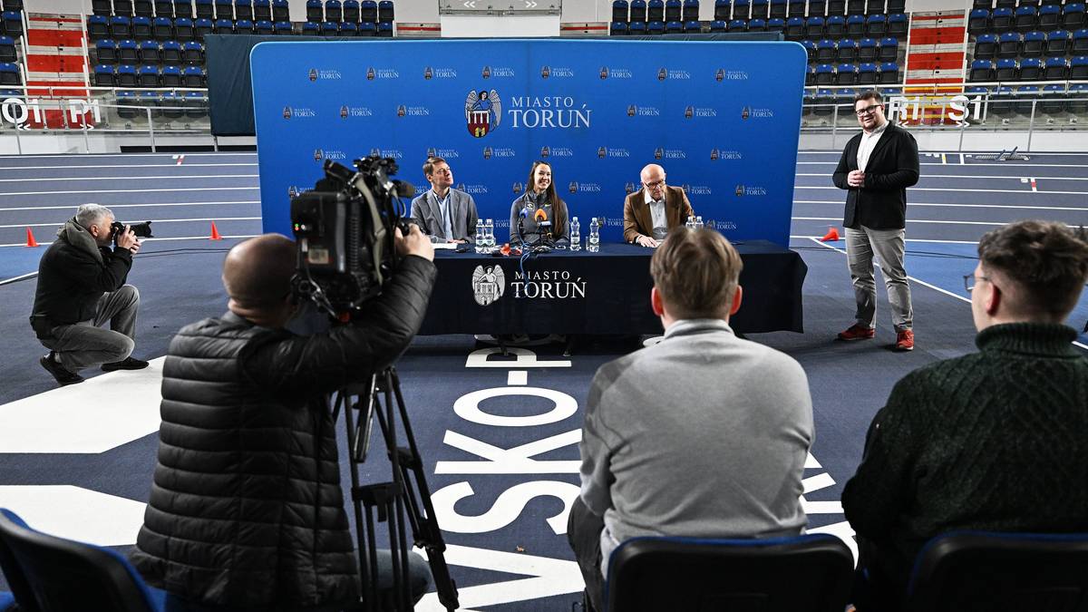 Konferencja prasowa przed ORLEN Copernicus Cup w Toruniu. Na scenie siedzą przedstawiciele organizatorów i sportowcy, za nimi widnieje niebieskie tło z logo wydarzenia. Wokół zgromadzeni są dziennikarze z kamerami i aparatami.