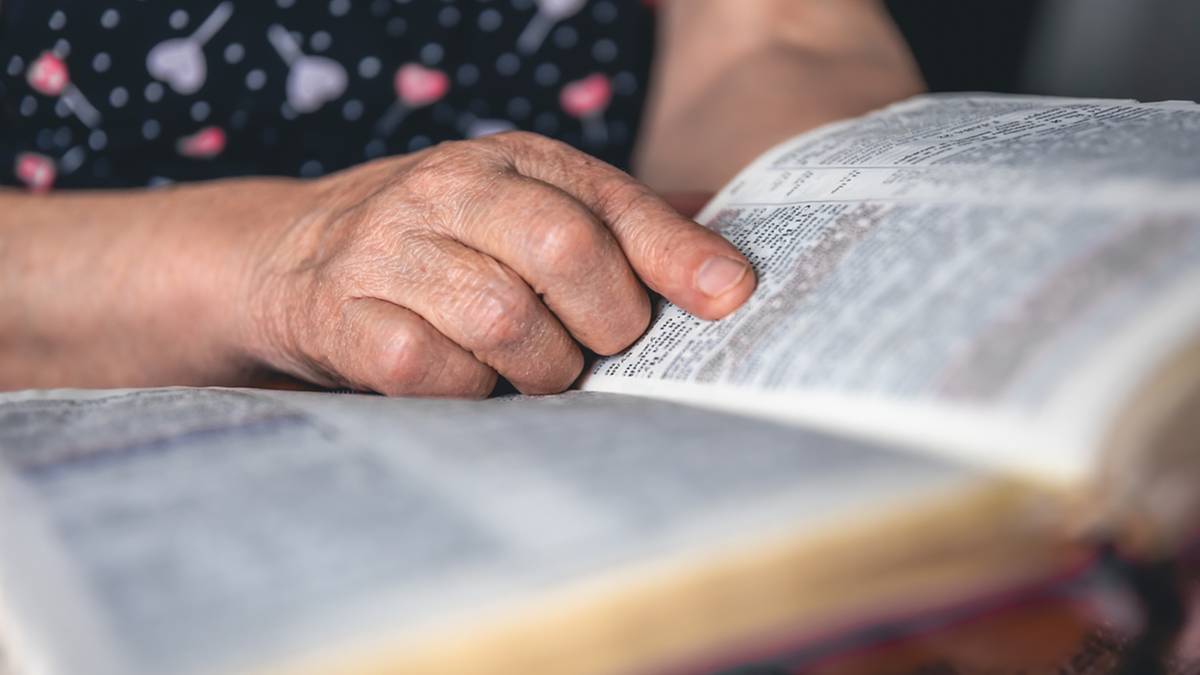 Dłoń starszej osoby dotykająca palcem otwartej książki z drobnym tekstem.