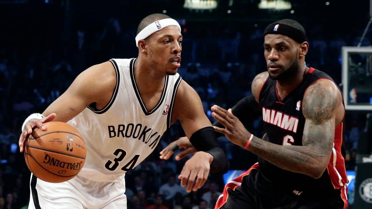 Dwóch zawodników NBA podczas meczu. Zawodnik w białej koszulce z napisem "BROOKLYN" i numerem 34, z opaską na głowie, drybluje piłką. Obok niego stoi zawodnik w czarnej koszulce z napisem "MIAMI" i numerem 6, z czarną opaską na głowie, próbując mu przeszkodzić.