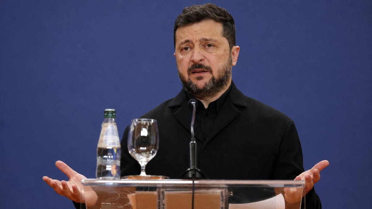 Wołodymyr Zełenski przemawia za m&oacute;wnicą z rozłożonymi rękoma na tle niebieskiej ściany.