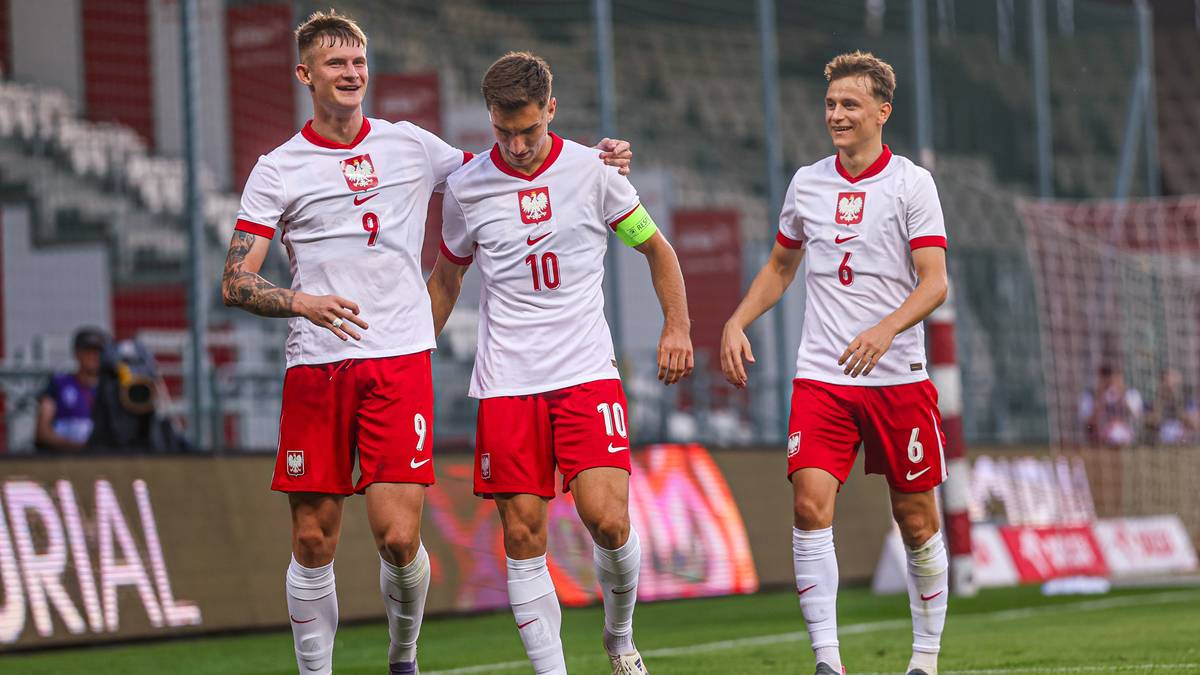 Trzech piłkarzy w białych koszulkach z czerwonymi elementami i czerwonych spodenkach, stoją obok siebie na boisku piłkarskim.