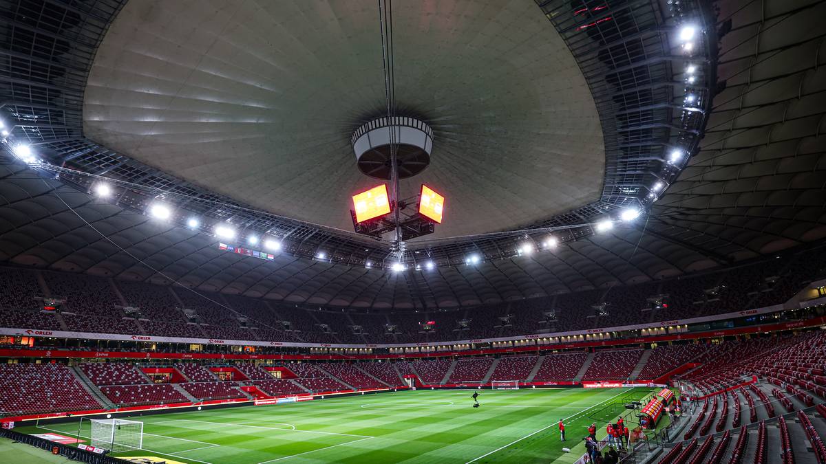 Widok na zieloną murawę stadionu piłkarskiego z oświetleniem i trybunami.