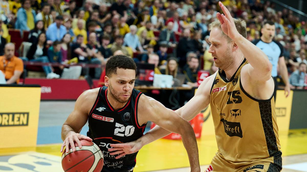 Dwóch zawodników drużyny koszykarskiej podczas meczu, jeden z nich prowadzi piłkę, drugi próbuje go zatrzymać. Widownia na trybunach w tle.