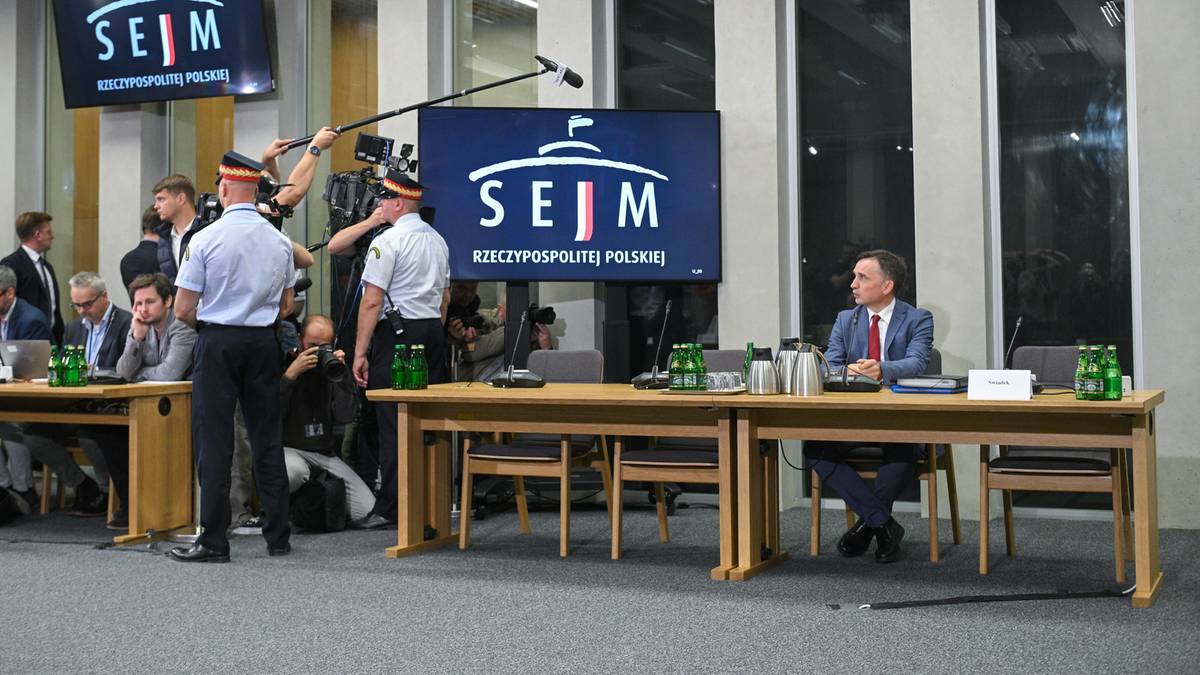 Sala sejmowa z dwoma ekranami wyświetlającymi logo Sejmu. Po prawej stronie siedzi mężczyzna w garniturze przy stole, a po lewej grupa osób i techników przygotowujących się do transmisji.