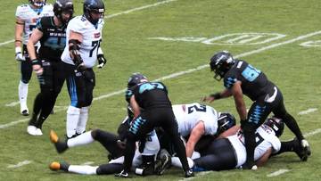 Drużyna w czarnych strojach z niebieskimi akcentami walczy o piłkę z drużyną w białych strojach z niebieskimi akcentami na boisku do futbolu amerykańskiego.