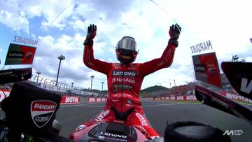 Motocyklista w czerwonym kombinezonie i kasku z uniesionymi rękami, na torze wyścigowym.