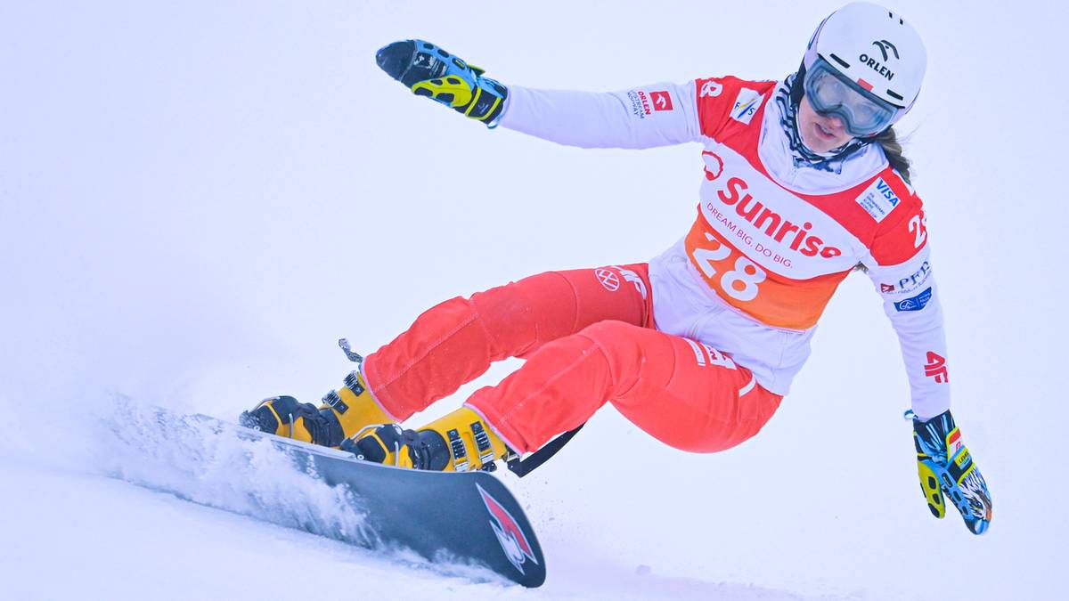 Snowboardzistka w kombinezonie z numerem 28 zjeżdża po stoku, robiąc zakręt.