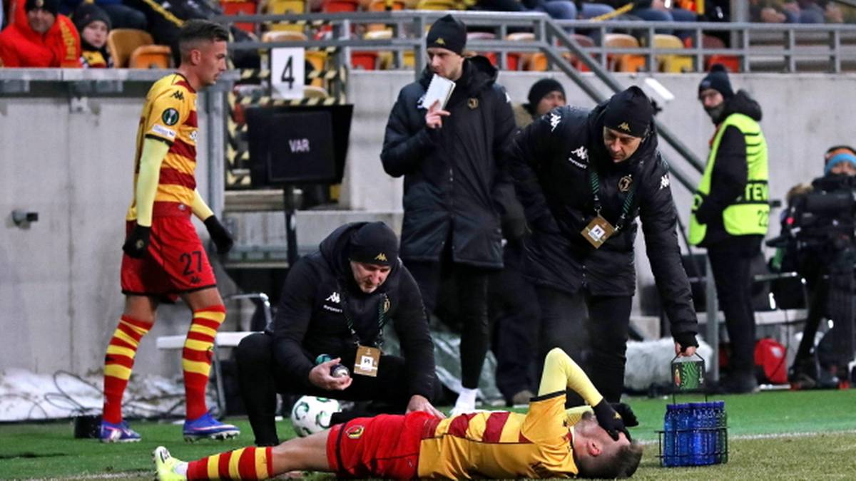 Zawodnicy Jagiellonii Białystok otaczają rannego kolegę z drużyny na boisku piłkarskim.