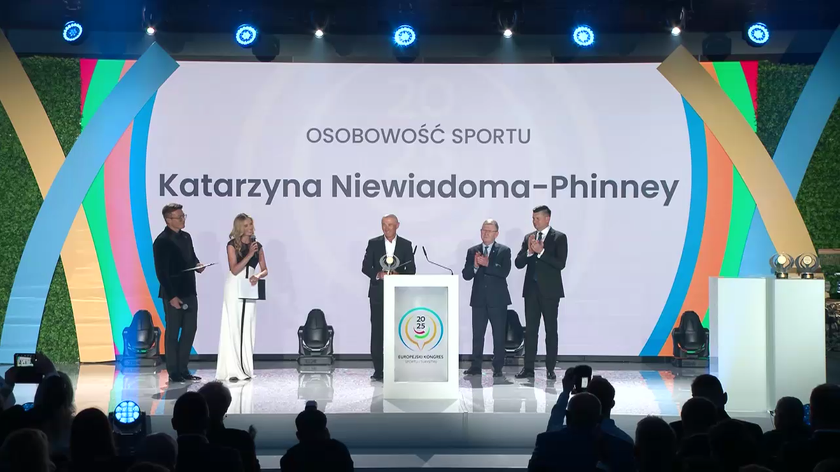 Kobieta i mężczyzna stojący na scenie przed podium z mikrofonem, wręczają statuetkę osobie stojącej za podium. Na ekranie za nimi widnieje napis "Katarzyna Niewiadoma-Phinney".