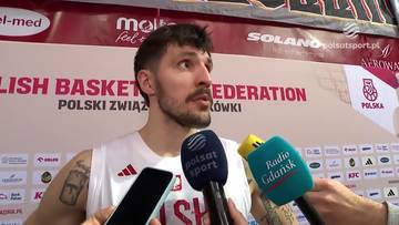 Dominik Olejniczak, koszykarz z brodą, ubrany w białą koszulkę z godłem Polski, odpowiada na pytania dziennikarzy przed kilkoma mikrofonami. W tle widoczny jest baner z napisem "POLISH BASKETBALL FEDERATION".