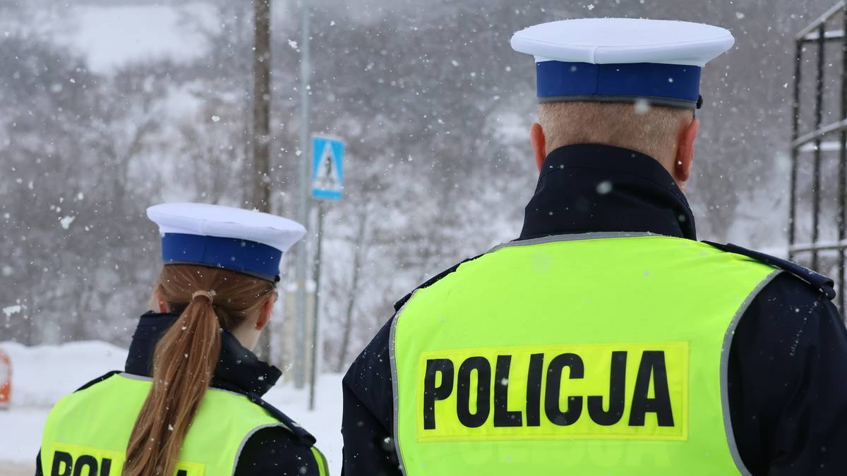 Dwóch policjantów w mundurach i kamizelkach odblaskowych z napisem POLICJA, stojących na tle ośnieżonego krajobrazu.